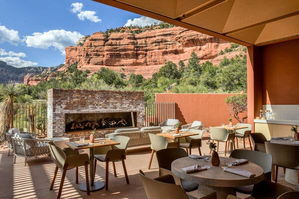 An outdoor dining area has intimate tables with sleek modern chairs, some placed within an open area with burnt orange walls and high ceilings. In the open area, a long outdoor fireplace is made of bricks; a couch and comfortable-looking chairs surround it. Beyond the terrace are the red cliffs and desert vegetation typical of Sedona, Ariz. Farther in the background are low mountains beneath a blue sky with puffy clouds.